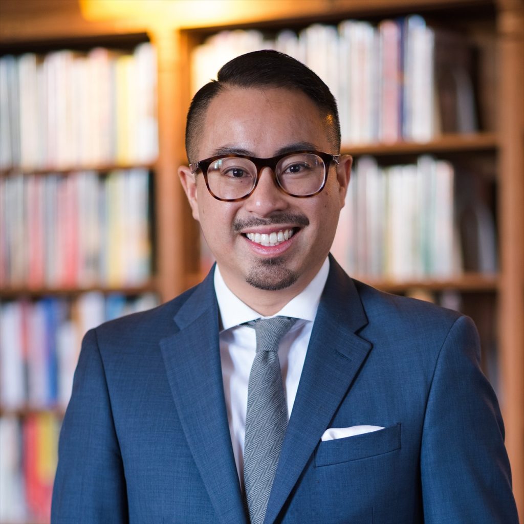 Photograph of Mike Hoa Nguyen in a blue suit and dark rimmed glasses with a bookshelf in the background