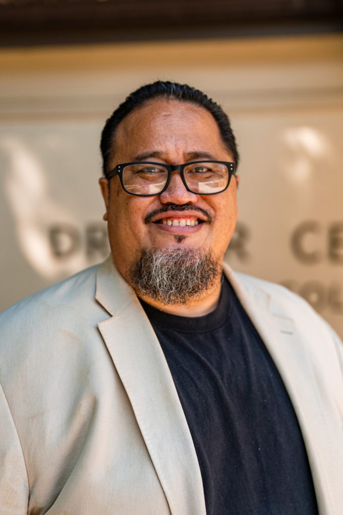 Photograph of Sefa Aina in a light colored suit jacket and black rimmed glasses
